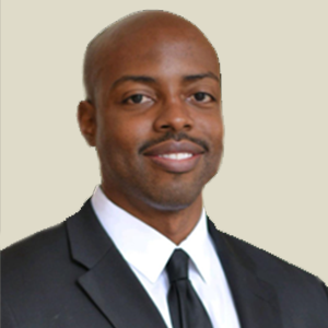 The image shows Evans St. Fort, a Board Member for 2023. He is smiling slightly, wearing a dark suit jacket, a white dress shirt, and a black tie. Evans has a bald head and a neat mustache. The light beige background highlights his professional and composed appearance.