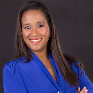 The image shows Lory M. Freiman, CPA, the Treasurer and a member of the board for 2023. She is smiling confidently with her arms crossed, wearing a bright blue blouse. Lory has long, straight hair, and the dark background contrasts with her attire, highlighting her poised and professional appearance.