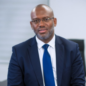 The image shows Carl Momplaisir, the Vice-Chairman and a member of the board for 2023. He is wearing a dark blue suit, a white dress shirt, and a blue tie. Carl has a neatly trimmed beard and is wearing glasses. His expression is serious yet approachable, and he is seated in a professional setting with a neutral background that complements his formal attire.