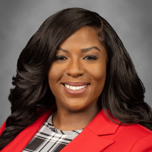 The image shows Dr. Sandra Severe, Ph.D., MPH, a Board Member for 2023. She is smiling confidently, wearing a red blazer over a black-and-white patterned top. Dr. Severe has long, wavy black hair, and the gray background complements her professional and polished appearance.