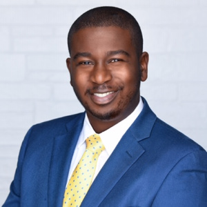 The image shows Vladimir Tranquille, a Financial Advisor and Board Member for 2023. He is smiling confidently, wearing a blue suit jacket, a white dress shirt, and a yellow tie with a subtle pattern. Vladimir has a short haircut, and the light-colored background with a brick-like texture complements his professional and approachable appearance.