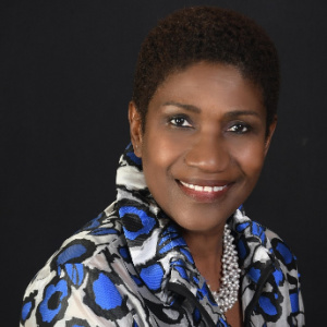 The image shows Gepsie M. Metellus, a Board Member for 2023. She is smiling warmly and is dressed in a patterned blouse with shades of blue, white, and black, paired with a pearl necklace. Gepsie has a short haircut, and the dark background emphasizes her elegant and professional appearance.