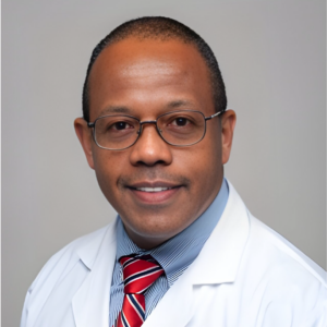 Portrait of Dr. Carmel J. Barrau in a lab coat, with glasses, a blue-striped shirt, and a red-striped tie, conveying a professional and knowledgeable demeanor.