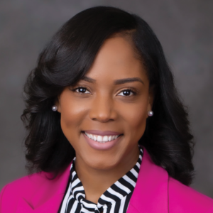 Portrait of Eurica Ketant, with styled waves in her hair, pearl earrings, a striped blouse, and a vibrant pink blazer, displaying a professional and cheerful demeanor.