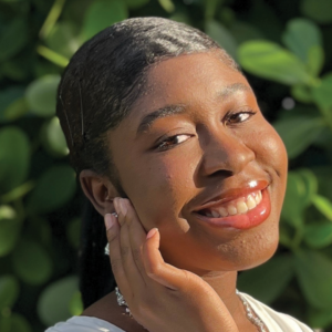 Close-up portrait of a smiling young woman named Gabriella Alexis, with glowing skin, wearing minimal makeup and a white outfit, accessorized with stud earrings and a delicate necklace. She is posing with her hand gently touching her cheek, against a lush green foliage backdrop.