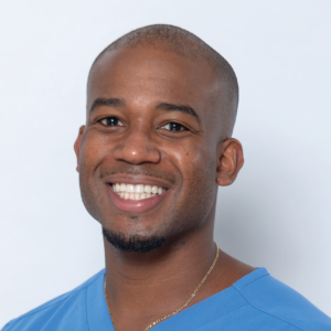 Portrait of Kyle Phanord with a bright smile, sporting a clean-shaven head, wearing a light blue shirt, and a gold chain necklace.