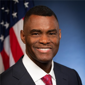 Official portrait of Markenzy Lapointe, Esq., with a confident smile, in a suit and tie, with an American flag in the background.