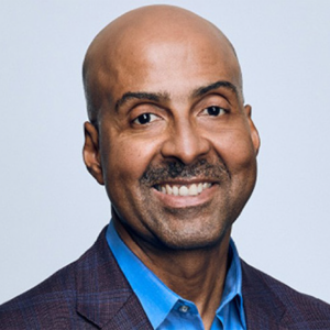 Portrait of Pascal DesRoches with a friendly smile, bald head, wearing a blue shirt and patterned blazer.