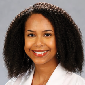 Professional headshot of a woman named Pascale Duvalsaint, with shoulder-length curly hair, a radiant smile, and wearing a white lab coat, indicating a healthcare or scientific profession.