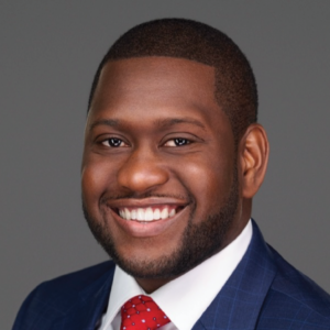 A professional headshot of Reno Pierre with a beaming smile, dressed in a navy blue suit with a red polka dot tie.