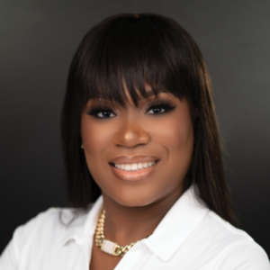 Headshot of Shavon Saint Preux with a cheerful smile, wearing a white shirt and a chunky gold necklace, her hair styled with bangs.