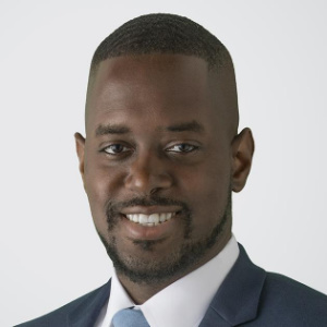 The image shows Pedro Gassant, Esq., a Board Member for 2023. He is smiling, wearing a dark suit jacket, a white dress shirt, and a light blue tie. Pedro has a short haircut and a neatly trimmed beard. The light background complements his professional and polished appearance.