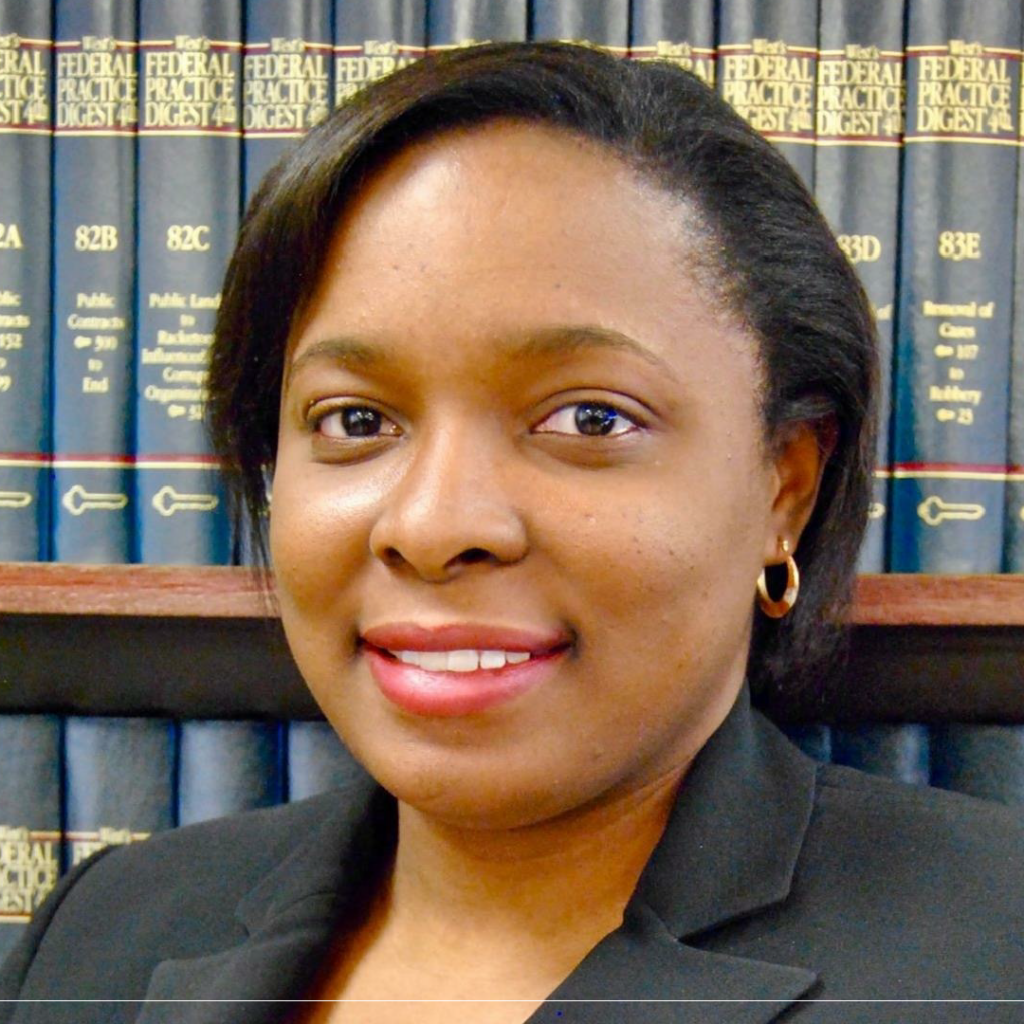 Portrait of Elisabeth Magloire, a professional woman with short black hair, wearing a black blazer and gold hoop earrings, standing in front of a bookshelf filled with legal books.