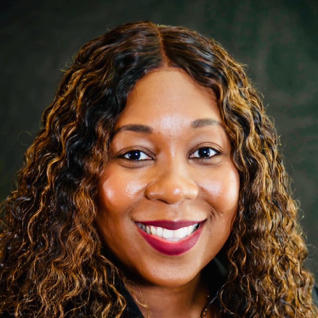 Headshot of Emmanuella Annette Telfort, smiling warmly. She has long, curly hair with highlights, and is wearing a dark outfit. The background is a soft, dark tone, emphasizing her friendly expression.