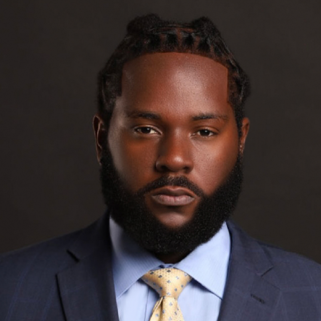 The image features a formal portrait of Mark St. Vil, a Haitian-American attorney. He is wearing a suit with a light blue dress shirt and a patterned tie. His hair is styled in neatly braided cornrows, and he has a full, well-groomed beard. Mark's expression is serious and professional against a dark, neutral background.