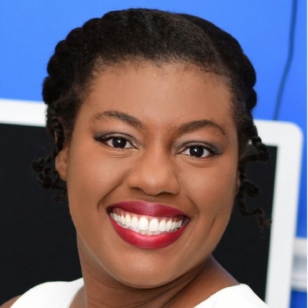 The image is of Nadeige Theresias-Joisil, an educator and community leader, smiling confidently. She has styled natural hair and is wearing red lipstick, conveying warmth and professionalism. The background features a blue backdrop and a computer screen, highlighting her role in a professional or educational setting.