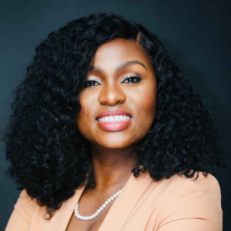 Portrait of Prisca Duclos, a woman with curly black hair, wearing a light brown blazer and a pearl necklace, smiling against a dark background.