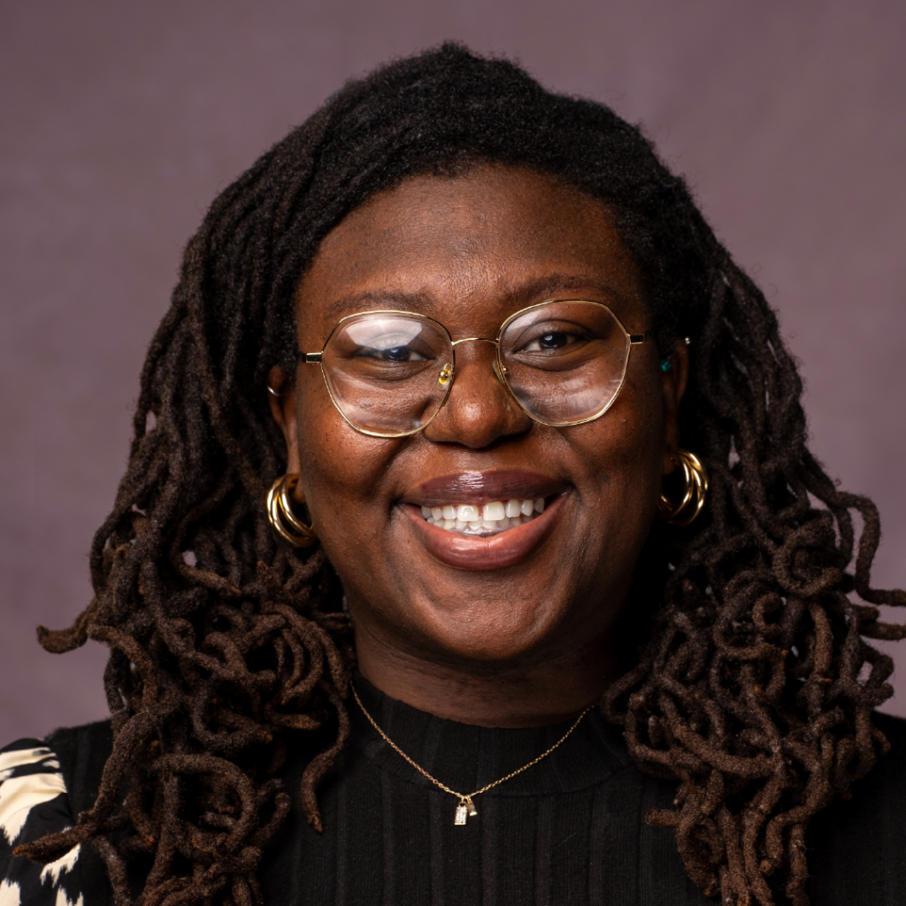 Headshot of Shanava Saintolien, a smiling Black woman with long locs, wearing glasses, gold hoop earrings, and a black top.