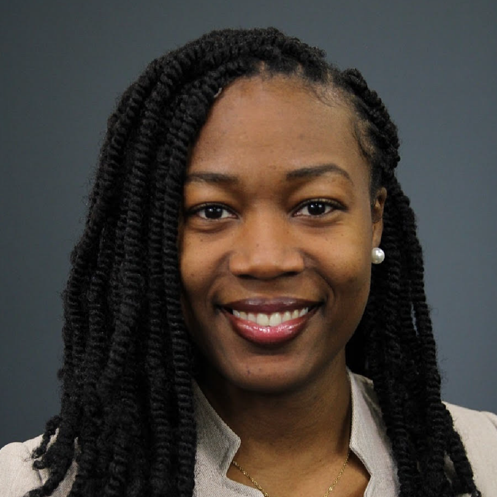 The image is a professional headshot of Dr. Vickie Dugat. She is smiling, with long, neatly twisted black hair. She is wearing pearl earrings and a light-colored blazer, set against a solid dark grey background. The image highlights her warm and approachable demeanor.