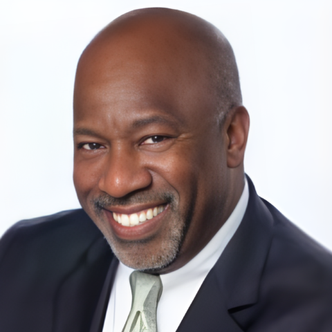 The image shows Jean G. Belizaire, the Vice-Chairman and a member of the board for 2023. He is smiling warmly, wearing a dark suit jacket, a white dress shirt, and a light-colored tie. Jean has a bald head and a neatly trimmed beard. The background is a light gradient, which complements his professional appearance and highlights his friendly demeanor.