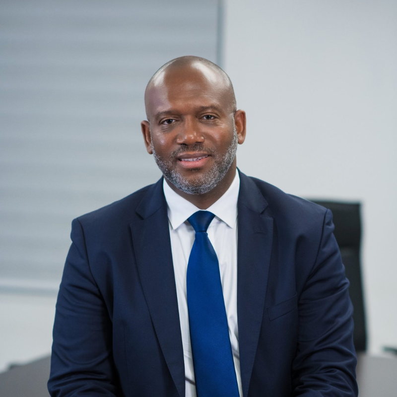 The image shows Carl Momplaisir, the Chairman and a member of the board. He is wearing a dark blue suit, a white dress shirt, and a blue tie. Carl has a neatly trimmed beard and is wearing glasses. His expression is serious yet approachable, and he is seated in a professional setting with a neutral background that complements his formal attire.